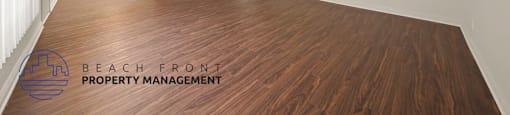 A spacious room with wooden floors and white walls, with a logo for Beach Front Property Management in the bottom left corner.