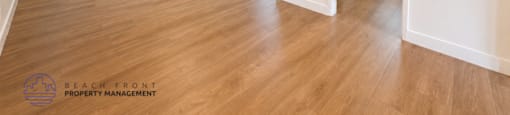 A spacious room with wooden flooring and white walls, with a logo for Beach Front Property Management in the bottom left corner.