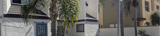 A white house with a black gate stands in front of a palm tree.