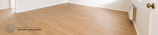 A room with wooden flooring and white walls, with a door on the left and a window on the right.