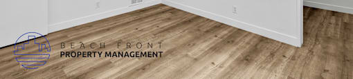 A room with wooden flooring and white walls, with a logo for Beach Front Property Management in the bottom left corner.