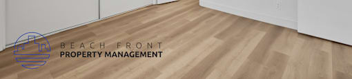 A room with white walls and wooden flooring, with a logo for Beach Front Property Management in the bottom left corner.