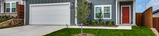 A grey house with a red door and a small tree in the front yard.