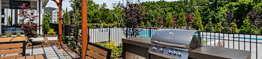 A wooden table and bench are on a patio with a grill and a pool in the background.