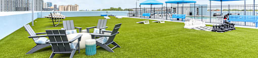A lawn with chairs and umbrellas is in the foreground with a pool and apartment buildings in the background.