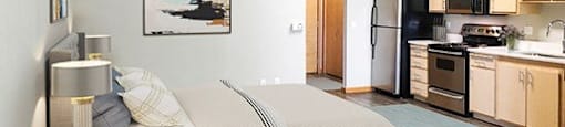 1 BR Apartments in Downtown Sacramento CA - Fremont Mews Apartments - Studio Bedroom Area with Wood-Style Flooring, Area Rugs, Bed, Nightstands with Lamps, Wall Art, and View of Kitchen at FREMONT MEW