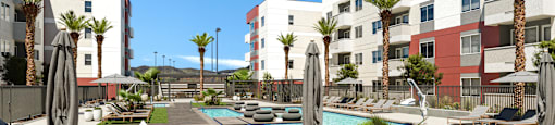 A pool surrounded by lounge chairs and umbrellas in front of apartment buildings.
