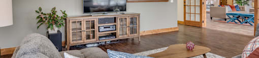Pet-Friendly Apartments In Spokane, WA - Deer Creek - Living Room With Grey Sectional Sofa, Wooden Media Console, And Open French Doors That Lead Into Another Sitting Area.