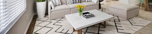 Apartments In Glendale, AZ, For Rent - Spectra West - Open-Concept Living Room With Wood-Look Flooring, Area Rug, Couch, Ottoman, Coffee Table, Houseplant, Large Window, And View Of Dining Area And Kitchen.