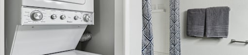A white washing machine sits next to a bathroom with a grey towel hanging on the wall.