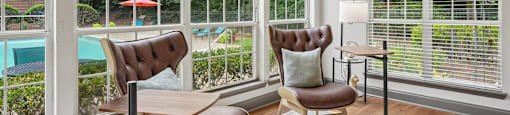 A room with a chair and a table in front of a window with a view of a pool and trees.
