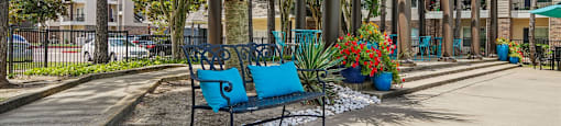 A blue bench sits in the middle of a concrete area with a fence and trees in the background.