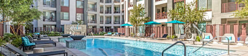 A pool surrounded by apartment buildings and lounge chairs.