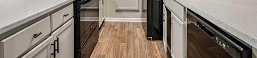 A black refrigerator stands in a kitchen with white cabinets and a wooden floor.