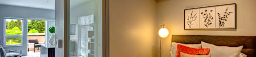 Bothell Apartments - 98th Avenue - A Bedroom With A Sliding Frosted Glass Door, Layered Pillows, Floor Lamp, And Wood-Style Flooring.