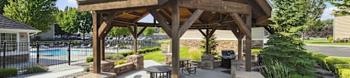 A wooden pavilion with a stone base is surrounded by greenery and a gravel path.