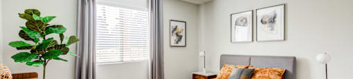 Senior Living Las Vegas, NV - Gramercy Parc - A Bedroom With Gray Wood-Style Plank Flooring, Gray Upholstered Headboard, Large Window With Blinds, Two Matching Wooden Nightstands, And A Plant.