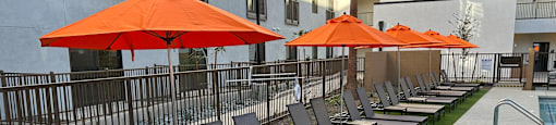 Studio Apartments in Downtown Phoenix, AZ - VB on 12th Street - Poolside Lounge Seating with Orange Umbrellas on a Patch of Artificial Grass Turf and Surrounded by a Fence.