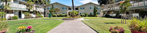 Apartments For Rent Gilroy, CA - Mission Park - Residential Courtyard In The Heart Of The Apartment Complex With Pebbled Paths, Potted Plants, Lush Green Grass, And Trees