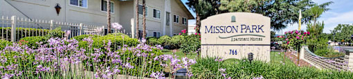 Apartments for Rent in Gilroy, CA - Mission Park - Exterior View of Property with Signage, Lush Landscaping, Walking Path, and Trees.