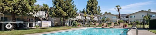 Apartment Gilroy, CA - Mission Park - Gated Sparkling Pool with Tan Lounge Chairs, Grassy Lawn, Trees, and View of Apartment Buildings in the Background.