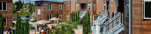Studios in Portland, OR - The Beverly - Courtyard with Umbrella Tables and Chairs, Bench, Potted Plants, Trees, and Surrounding Apartment Buildings.