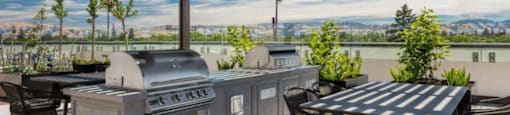Santa Clara Apartments - Santana Terrace -  A Grilling Area with a Covered Pergola, Stainless-Steel Grills, Potted Plants, and Dining Tables.