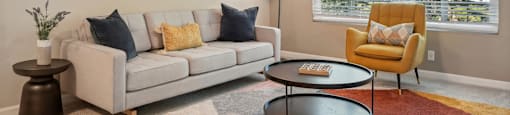 Two-Bedroom Apartments in Fremont, CA - Northgate Savoy - Furnished Living Room with Carpeted Flooring, Cream Couch, Yellow Armchair, Wooden Side Table, Standing Lamp, Round Brown Coffee Table, Large Window, Ceiling Fan, and Wall Art.