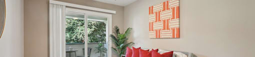 One-Bedroom Apartments in Fremont, CA - Northgate Savoy - Bedroom with Carpeted Flooring, Bed, Wooden Nightstand, Houseplant, Ceiling Fan, Beige Walls, Framed Mounted Mirror, and Sliding Glass Door to Balcony.