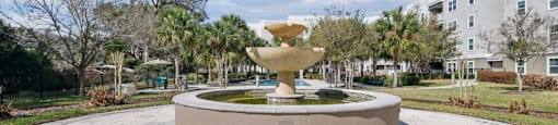 A fountain in the middle of a paved area with trees and buildings in the background.