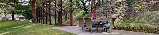 Daly City, CA, Apartments - Serra Commons - Picnic Area with Concrete Flooring, Table, Chairs, Manicured Lawn, and Surrounding Trees.