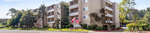 Apartments in Daly City, CA, for Rent - Serra Commons - Street View of Apartment Building with Property Signage, Flags, and Trees.