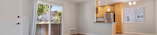 Dog-Friendly Apartments in Milpitas, CA - Spring Valley - Unfurnished Living Room Space with Wood-Style Flooring, Sliding Glass Door to Patio, White Walls, AC Unit, and View of Kitchen.