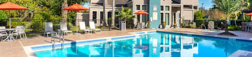 Pet-Friendly Apartments in League City, TX - Anchor at South Shore - Blue Outdoor Pool with a Neaby Hot Tub, Orange Umbrellas, Lounge Chairs, and Surrounding Greenery