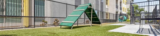 A playground with a green slide and a green bench in front of a grey building.