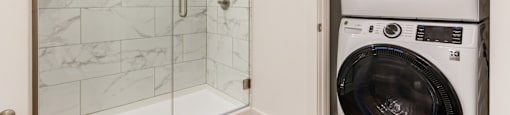 A white bathroom with a wooden floor, a marble shower, and a in-unit dryer/washer.