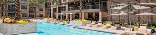 A large swimming pool surrounded by lounge chairs and umbrellas in front of a multi-story apartment building at 27Seventy Lower Heights apartments in Houston, TX.