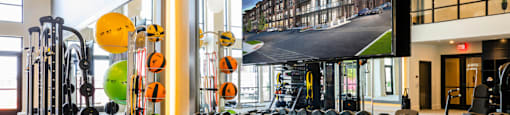 A gym with a variety of equipment including dumbbells, weight plates, and a television screen showing a building at 27Seventy Lower Heights apartments in Houston, TX.