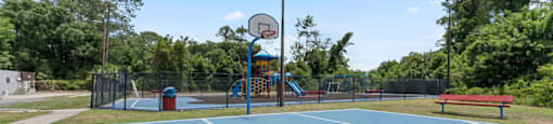 A basketball court with a blue surface and white lines, surrounded by trees and a red bench.