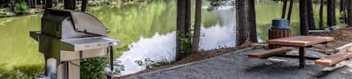 A picnic area with a grill and picnic table by a lake.