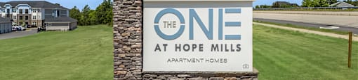 A sign for The One at Hope Mills Apartment Homes sits in a grassy field.