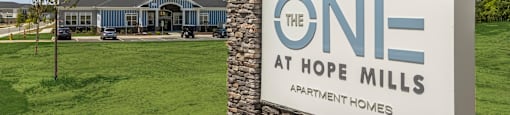 A sign for The One at Hope Mills Apartment Homes stands in front of a grassy field.