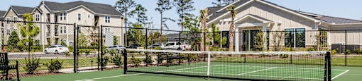 A tennis court is surrounded by a fence and a building. at Evolve Waterford Apartments in Belville, NC