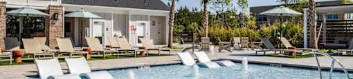 A pool with a waterfall and a patio with chairs and umbrellas at Evolve Waterford Apartments in Belville, NC
