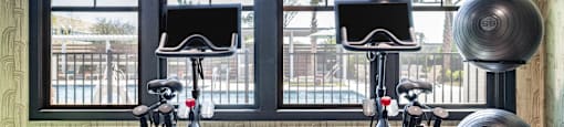 Two stationary bikes are in a room with a large window. at Evolve Waterford Apartments in Belville, NC