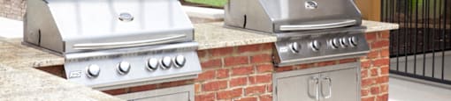 Outdoor grilling station at 1287 Shoals Apartments in Athens, GA, featuring two stainless steel gas grills built into a brick countertop, with a view of the landscaped grounds and residential buildings.