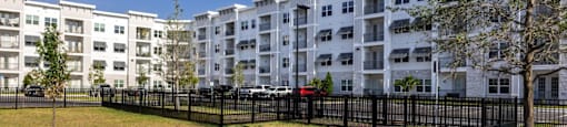 A row of white apartment buildings with black fencing and green trees in front.