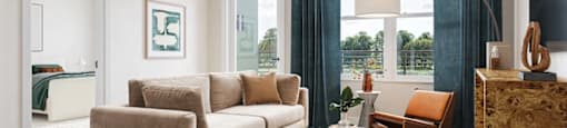 A living room with a beige sofa, a grey coffee table, and a large window with blue curtains at Boardwalk at Research Park apartments in Huntsville, AL.