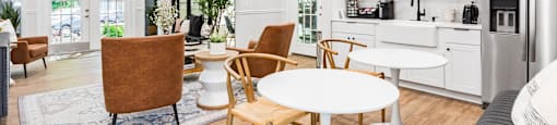 a living room with tables and chairs and a kitchen with white cabinets