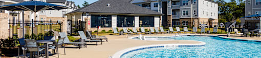A swimming pool surrounded by lounge chairs and umbrellas in front of apartment buildings.